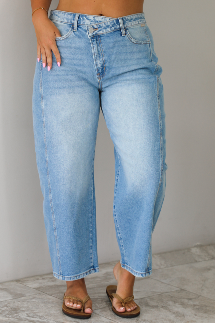 A woman wearing light-wash blue cropped barrel cut jeans with an asymmetrical button closure, standing indoors against white wall with white marble flooring. 