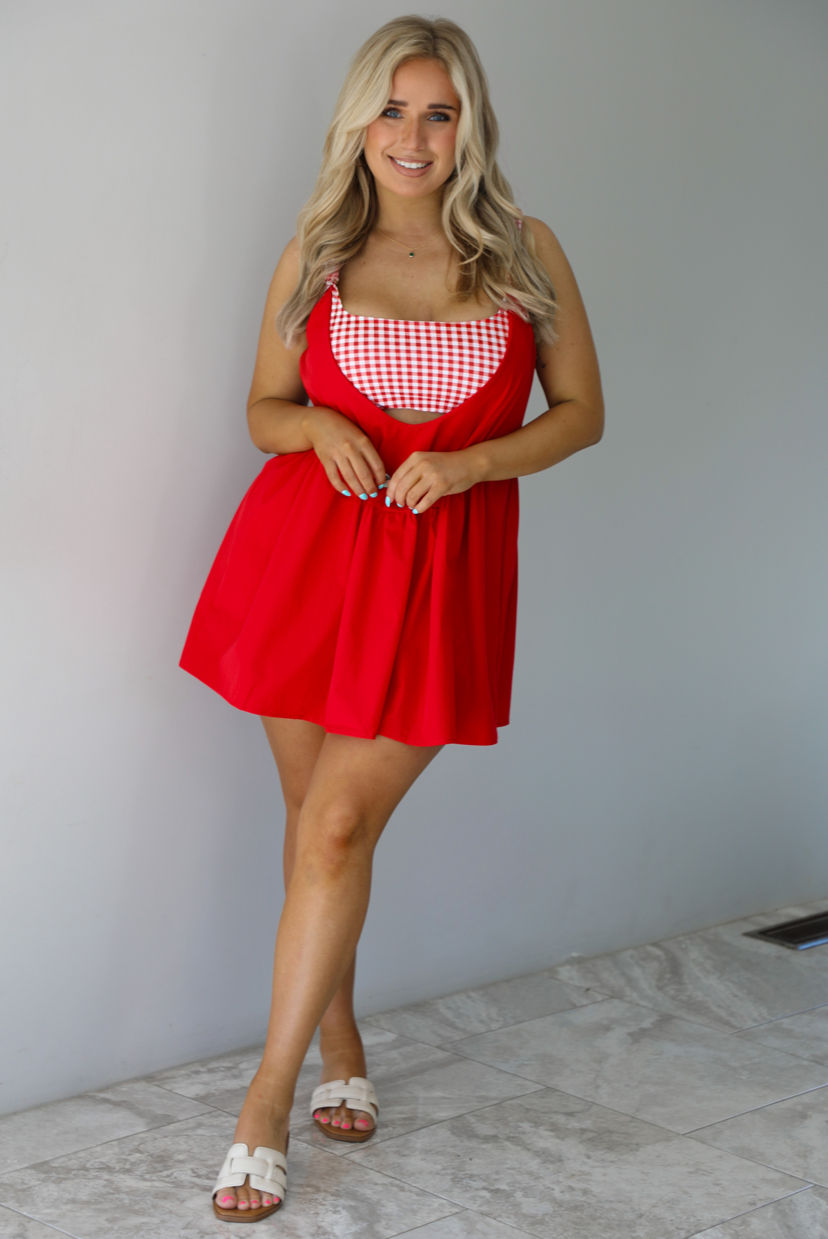 Woman wearing a red dress with a red and white gingham romper underneath. The photo is taken indoors against a white wall with white marble flooring. 