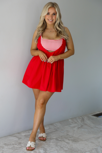 Woman wearing a red dress with a red and white gingham romper underneath. The photo is taken indoors against a white wall with white marble flooring. 