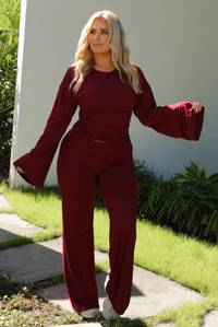 Woman model wearing a burgundy wine colored two piece set outdoors. The set features a slightly cropped, long sleeved top that has flared sleeves and a boat neckline. The pants are a mid-rise, fitted on the thighs, and have a straight cut design at the bottom. It's super soft, lightweight, and stretchy. 