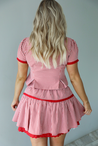 Woman wearing a red and white gingham patterned set that has a short sleeved button-up top that has pleated details at the hem of the top and a high waisted skirt with red lace detailing at the hem. The photo shows a closeup of the back of the set. The photo is taken indoors against a white wall.