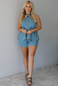 Woman wearing denim blue with white polka dot halter tie top and high waisted shorts indoors against a white wall with white marble flooring. 