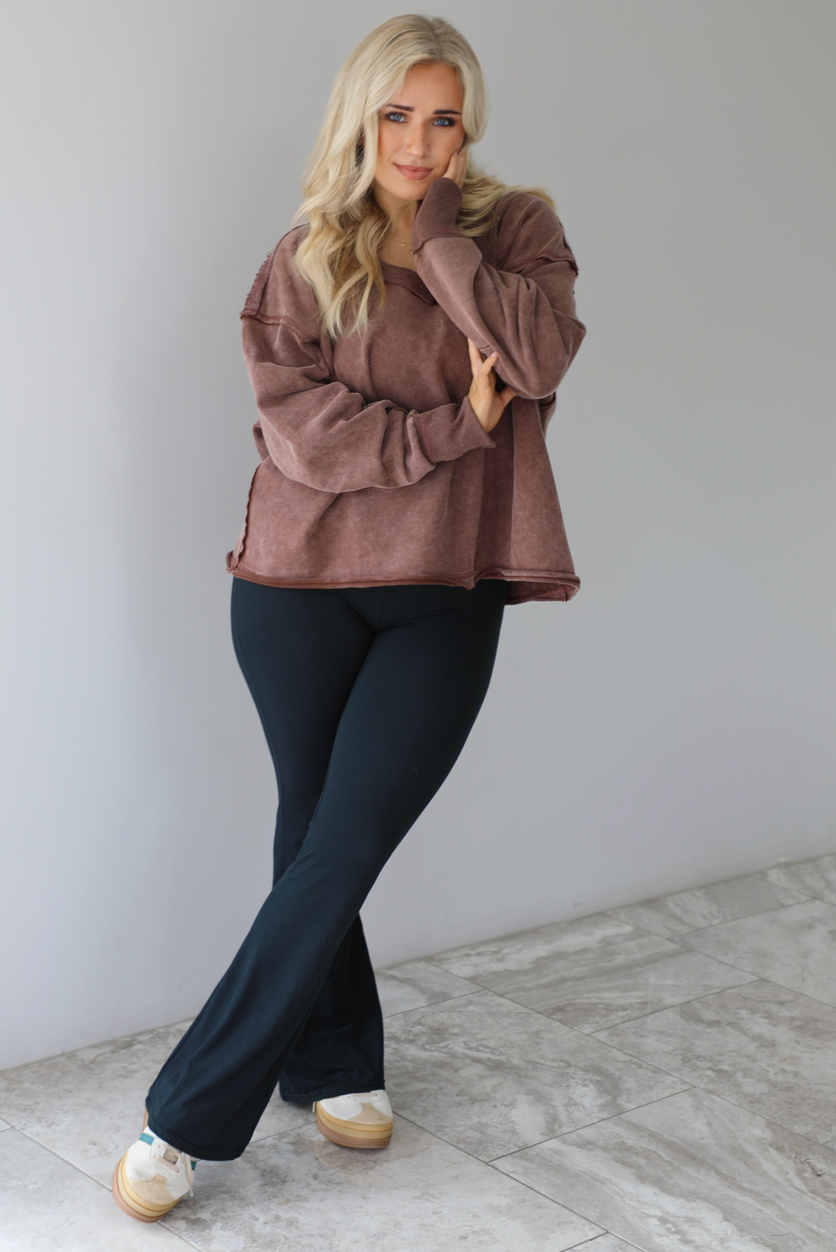 Woman wearing a mauve oversized v-neck sweater with raw hem details with high waisted black flared leggings. The photo is taken in front of a white wall with white marble flooring. 