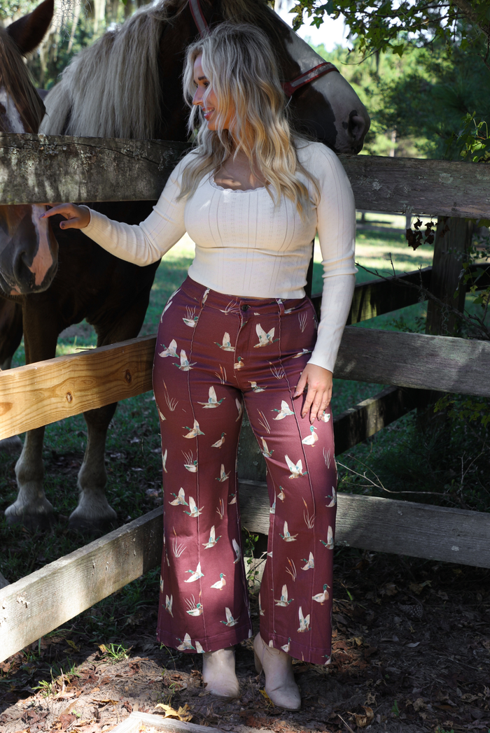 A woman wearing a cream colored knit long sleeved top paired with high waisted maroon jeans with a repeating mallard design, standing outdoors in front of a wooden fence and horses in the background. 