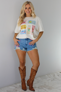 Woman wearing a white relaxed fit graphic tee that has various tickets on it with different fruits and flowers on them. The tee is tucked into a pair of distressed denim shorts and worn with a pair of cognac cowboy boots. The photo is taken indoors against a white wall with white marble flooring. 
