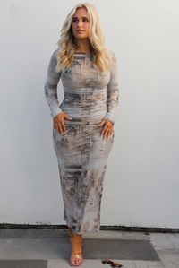 Woman model wearing a multi-colored and multi-patterned maxi length dress that is fitted with long sleeves. The photo is taken outdoors against a white wall. 