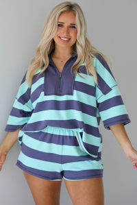 Woman wearing a turquoise and navy thick striped set featuring a collared short sleeved quarter zip top and high waisted shorts that have an elastic drawstring waistline with functional pockets on either side. The photo is taken indoors against a white wall. The photo shows a closeup of the set. 