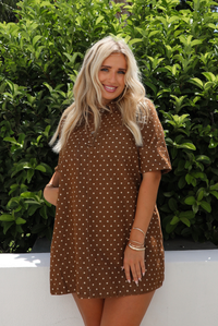 A woman with blonde hair wearing a brown short-sleeve mini dress with a cream polka-dot pattern, standing outdoors in front of a green hedge.