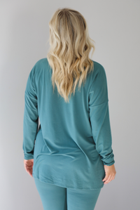 Woman wearing a teal two piece set that features an oversized long sleeved top and fitted leggings. The photo shows a closeup of the back of the top. The photo is taken indoors against a white wall.