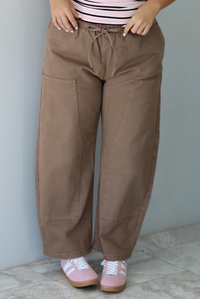 A closeup of a woman wearing mocha barrel pants that have an elastic and drawstring waistline, paired with a pink, burgundy, and white striped design and pink sneakers, standing indoors against a white wall with white marble flooring. 