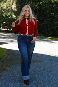 Woman model wearing a red cardigan with sand trim details and black button closures in the front with dark wash denim. The photo is taken outdoors on gravel with greenery in the back. 