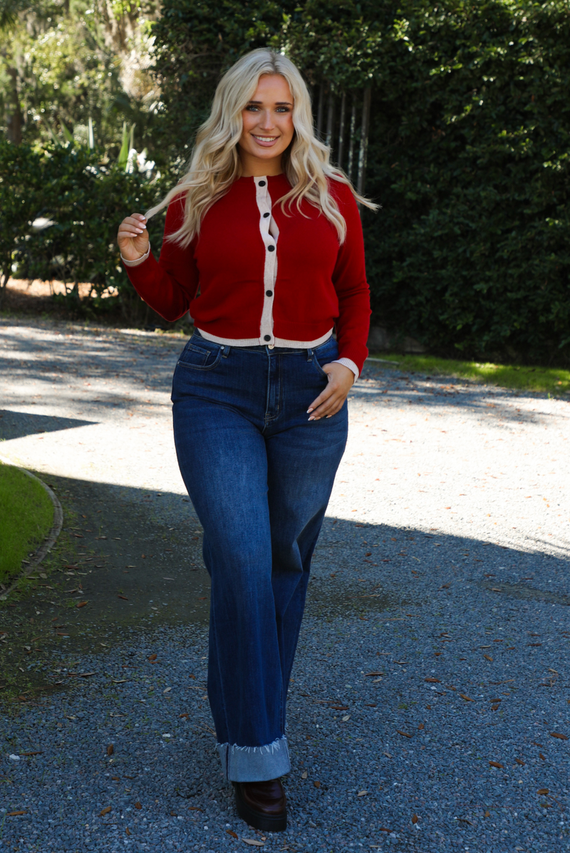 Woman model wearing a red cardigan with sand trim details and black button closures in the front with dark wash denim. The photo is taken outdoors on gravel with greenery in the back. 