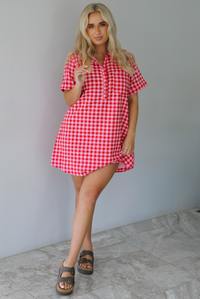 Woman wearing a pink and red gingham collared neckline t-shirt dress indoors against a white wall with white marble flooring. 