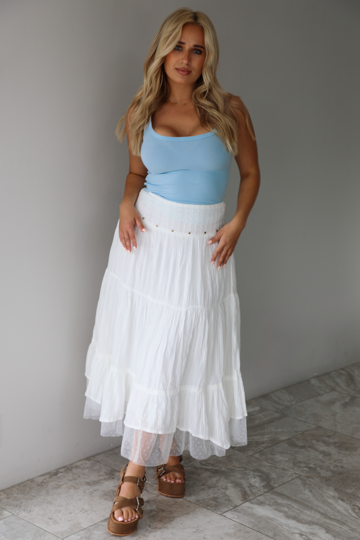Woman wearing a blue tank and high waisted white maxi-length skirt indoors against a white wall with white marble flooring. 