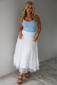 Woman wearing a blue tank and high waisted white maxi-length skirt indoors against a white wall with white marble flooring. 