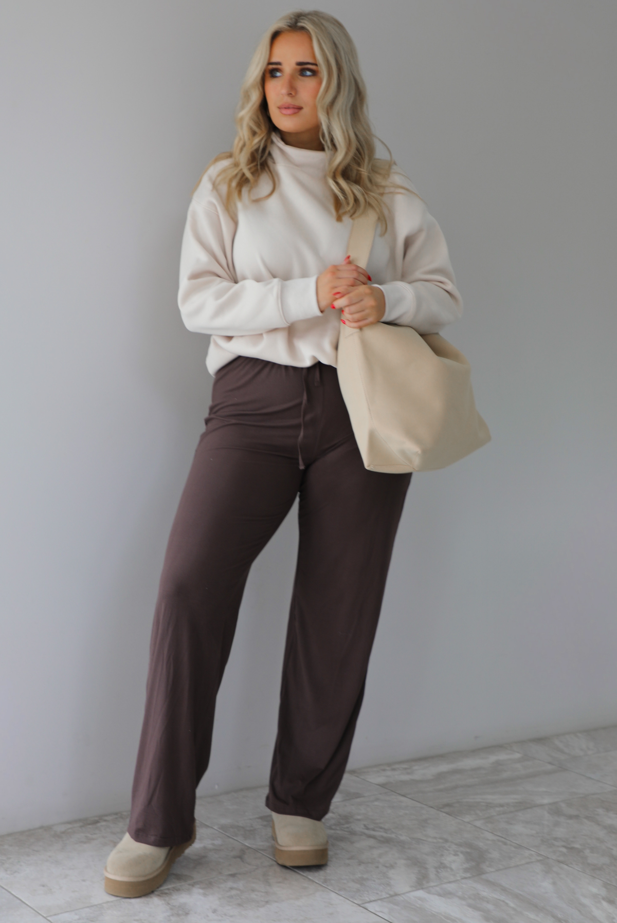 Woman wearing a beige high-neck, oversized sweater, high waisted brown pants, with a beige shoulder bag, and beige ugg boot shoes. The photo is taken indoors against a white wall with white marbled flooring. 