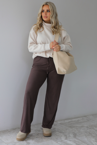 Woman wearing a beige high-neck, oversized sweater, high waisted brown pants, with a beige shoulder bag, and beige ugg boot shoes. The photo is taken indoors against a white wall with white marbled flooring. 