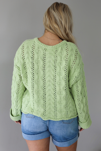 Woman wearing a green, long sleeved sweater with a loose knit design, functional button closures in the front, cuffed sleeves, and a raw, unfinished hem with medium wash denim shorts indoors against a white wall. The photo shows the back of the sweater. 