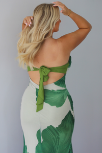 A blonde woman shown from behind models a strapless, bodycon maxi dress with a green tropical leaf print on a light beige background, with a tie closure in the back, standing indoors against a white wall.