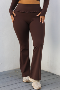 Woman model wearing a brown two piece athleisure set. The photo showcases the high waisted pants that have a ruffled waistline and a flared hem. The photo is taken outdoors against a white wall. 