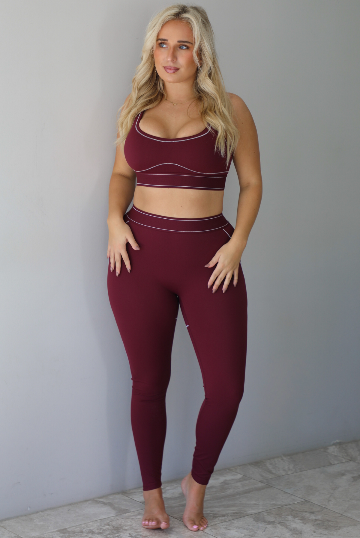 A woman wearing a maroon matching two piece set featuring a sports bra and high waisted leggings with white stitching details, standing indoors against a white wall with white marble flooring. 