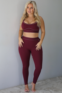A woman wearing a maroon matching two piece set featuring a sports bra and high waisted leggings with white stitching details, standing indoors against a white wall with white marble flooring. 
