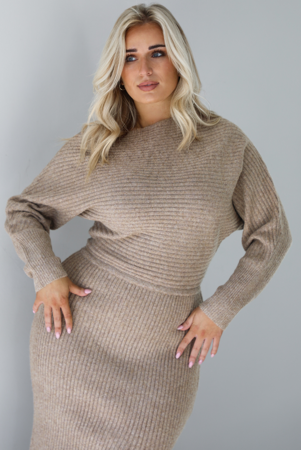 Woman wearing an oatmeal colored sweater dress that has a slouchy bodice that has long sleeves, with a fitted midi length skirt. The dress has a ribbed textured material that's horizontal on the bodice and vertical on the skirt. The photo is taken in front of a white wall.