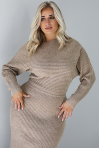 Woman wearing an oatmeal colored sweater dress that has a slouchy bodice that has long sleeves, with a fitted midi length skirt. The dress has a ribbed textured material that's horizontal on the bodice and vertical on the skirt. The photo is taken in front of a white wall.