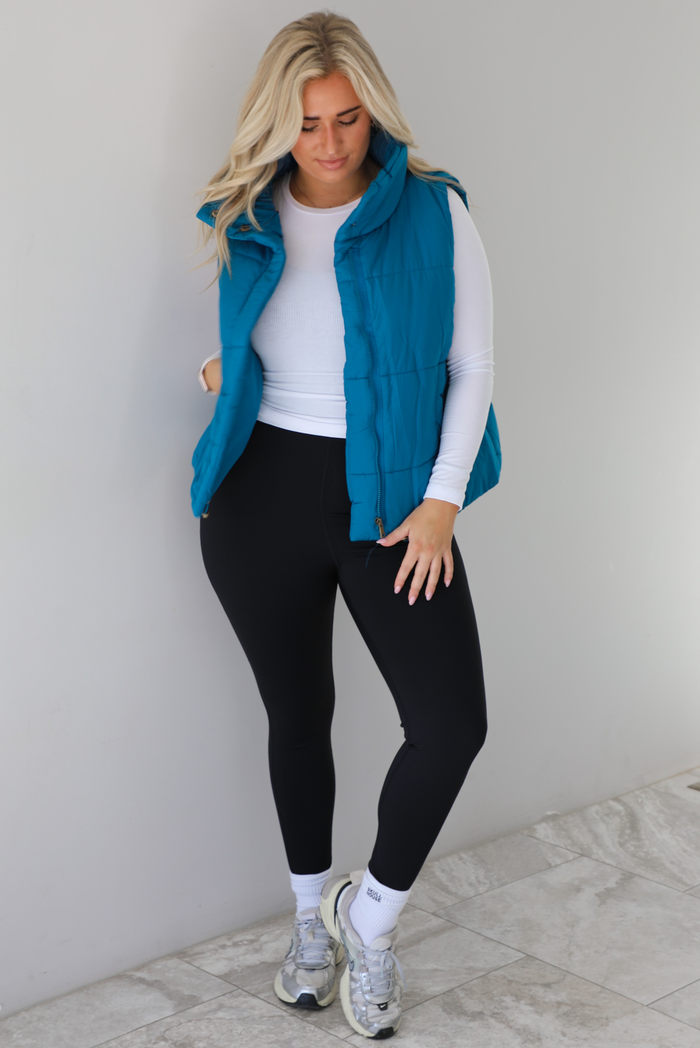 Woman wearing a teal puffer vest, white long-sleeve shirt, black leggings, and gray sneakers with white ankle socks indoors against a white wall with white marble flooring.