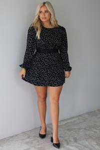 A woman wearing a black mini dress with a white polka dot print pattern throughout that has long sleeves, a high neckline, and a flowy skirt, standing indoors against a white wall with white marble flooring. 