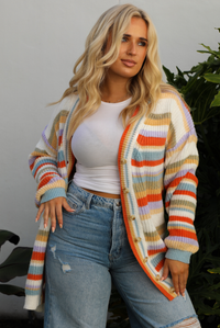 Woman model wearing an orange, lavender, yellow, muted green, blue, and white striped oversized cardigan with functional button closures in the front and an oversized fit with a light wash flared hem denim. The photo is taken outdoors against a white wall and greenery in the back. The photo showcases a close up of the cardigan unbuttoned and shows the full length, which falls to the upper thigh on our 5'7" model.
