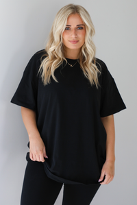 Woman wearing a black two piece set featuring an oversized t-shirt and fitted leggings. The photo shows a closeup of the oversized top. The photo is taken indoors against a white wall.
