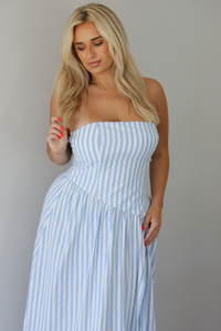 Woman wearing a blue and white striped strapless maxi dress indoors against a white wall. The photo shows a closeup of the bodice of the dress, showing how it has an asymmetrical drop waist. 