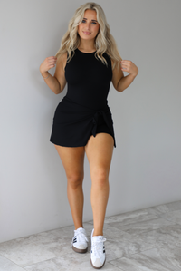 Woman wearing a black athleisure romper that has a wrap-style skirt detail that wraps around and ties in the front on the left side. It's styled with white sneakers that have black stripes down the side. The photo is taken indoors against a white wall with white marbled flooring. 