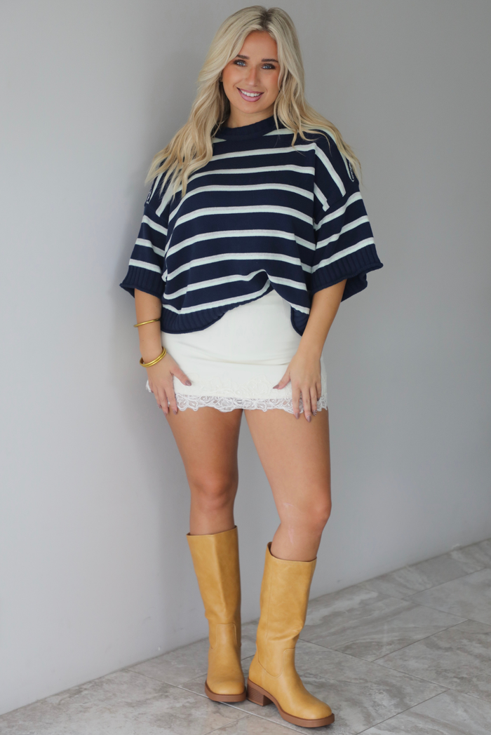 A woman wearing a navy and multi-colored striped design knit top paired with a fitted cream, lace trim mini skirt, standing indoors against a white wall with white marble flooring. 