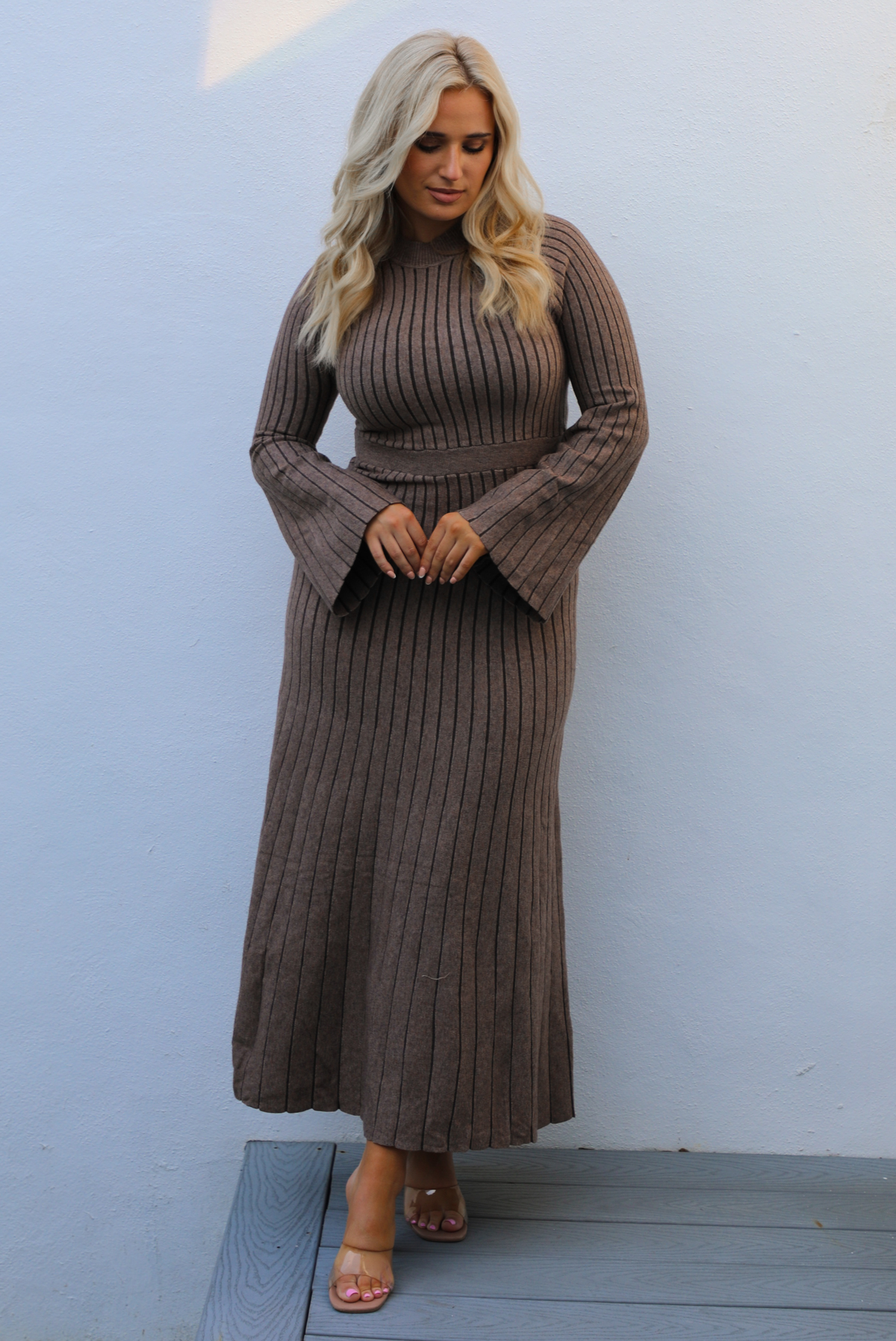 Woman wearing a taupe colored dress that has a vertical ribbed pattern throughout, a high neckline, fitted bodice, cinched waistline, long sleeves that flare slightly, and a maxi length skirt that flows at the bottom. The photo is taken outdoors against a white wall. 