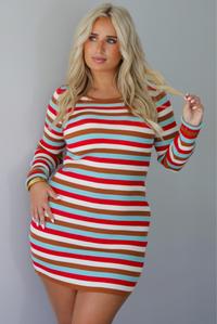 A woman wearing a red, blue, white, and brown striped long sleeved mini dress, standing indoors against a white wall.