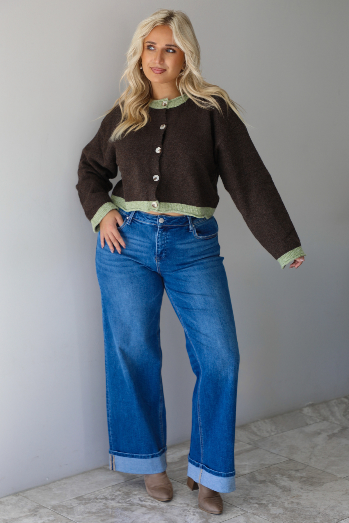 A woman wearing a dark brown with green trim cardigan fully buttoned up paired with dark wash, cuffed hem denim jeans, standing indoors against a white wall with white marble flooring. 