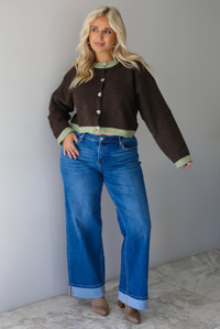 A woman wearing a dark brown with green trim cardigan fully buttoned up paired with dark wash, cuffed hem denim jeans, standing indoors against a white wall with white marble flooring. 