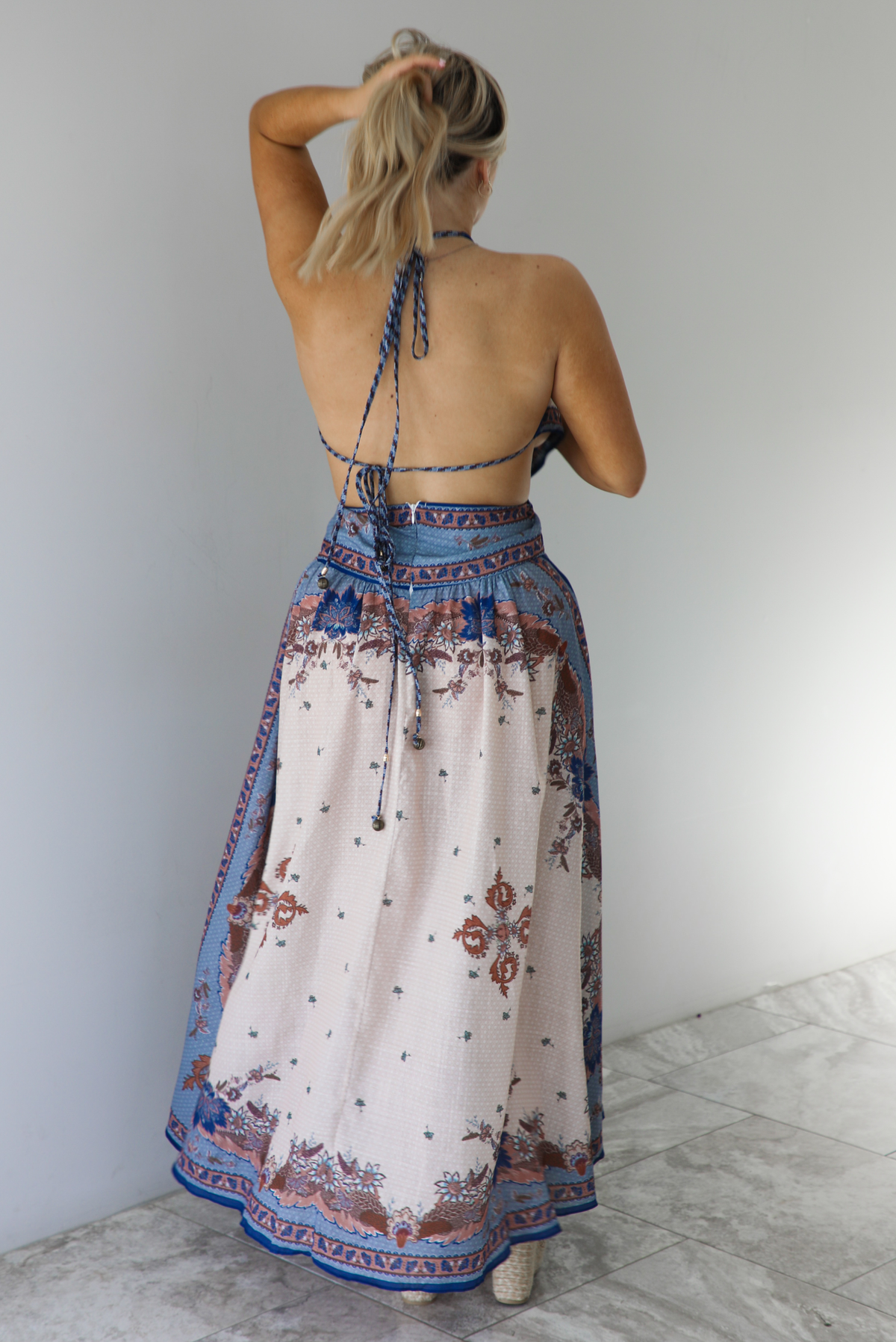 Woman wearing a blue and multi-colored halter tie maxi dress that has a long, flowy skirt with a split hem detail. The photo is taken indoors against a white wall with white marble flooring and shows the back of the dress, which has two tie closures, and a zipper closure. 