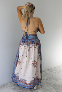 Woman wearing a blue and multi-colored halter tie maxi dress that has a long, flowy skirt with a split hem detail. The photo is taken indoors against a white wall with white marble flooring and shows the back of the dress, which has two tie closures, and a zipper closure. 