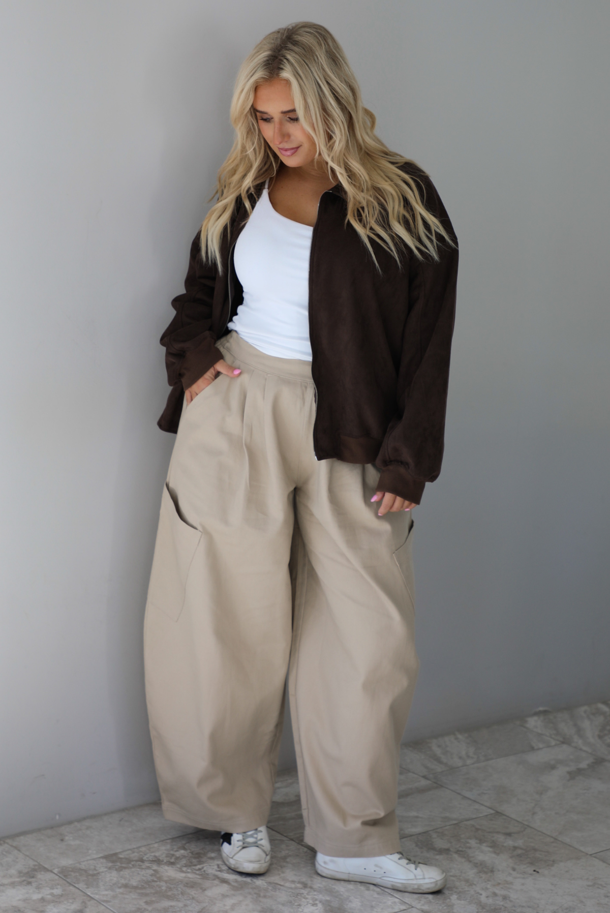 A woman wearing a brown suede jacket with a white tank and sand colored baggy pants, standing indoors against a white wall with white marble flooring. 