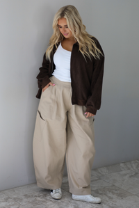A woman wearing a brown suede jacket with a white tank and sand colored baggy pants, standing indoors against a white wall with white marble flooring. 