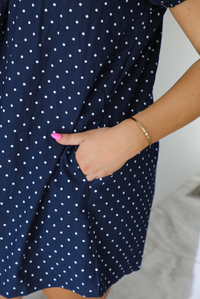 Woman wearing a navy baby-doll cut dress with a white polkadot print pattern throughout. The dress has puffed short sleeves and functional pockets on either side.  The photo shows a closeup of the pocket. The photo is taken indoors against a white wall.