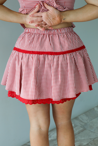 Woman wearing a red and white gingham patterned set that has a short sleeved button-up top that has pleated details at the hem of the top and a high waisted skirt with red lace detailing at the hem. The photo shows a closeup of the back of the skirt. The photo is taken indoors against a white wall.