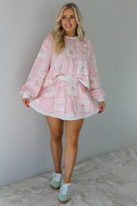 Woman wearing a two piece set featuring a pink floral set that has an oversized long sleeved top and high waisted skirt. The photo is taken indoors against a white wall with white marble flooring. 