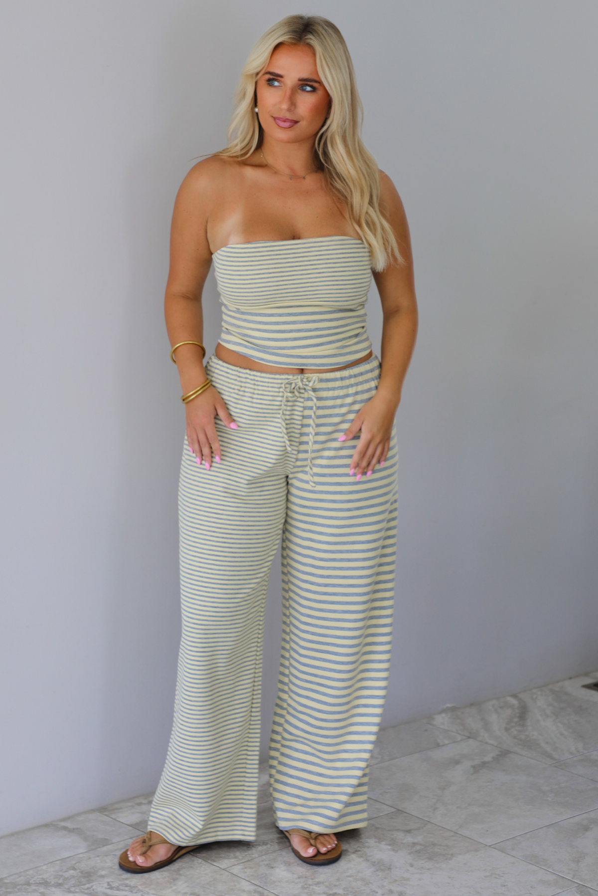 A woman wearing a pale yellow and heather grey striped two piece set with a sleeveless tube top and wide-leg pants, standing indoors against a white wall with white marble flooring. 