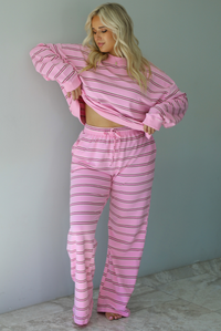 A woman wearing a pink two piece sweat set featuring a maroon and white striped design on a sweatshirt and high waisted, wide leg pants, standing indoors against a white wall with white marble flooring. 