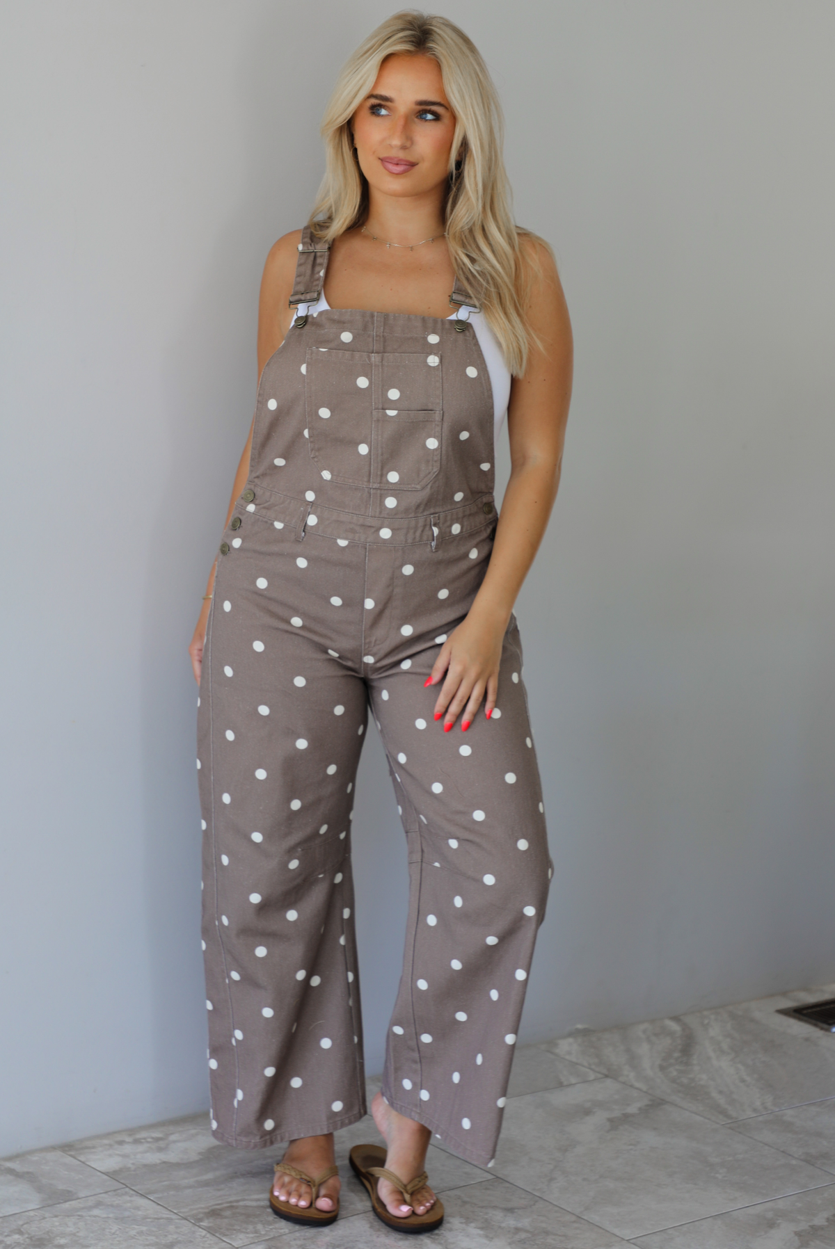 Woman wearing a brown with white polka dots overalls jumpsuit indoors against a white wall with white marble flooring. 