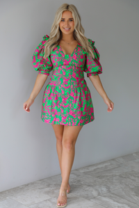 Woman wearing a fuchsia and green dress that has puffed sleeves, a v-neckline, fitted bodice, and flared skirt with pleated details indoors against a white wall with white marble flooring. 